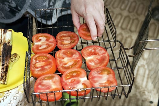 laying of fresh sliced tomatoes on the grate for roasting, visible hands of the cook, cooking for the picnic