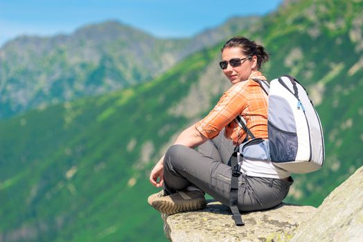 portrait of a woman sitting on a rock in the mountains with a backpack, relaxing with beautiful scenery