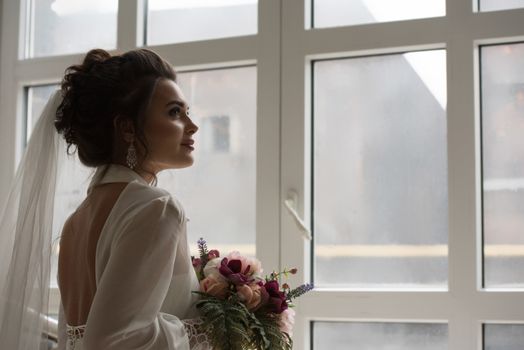 Beautiful bride in her wedding day with flower bouquet looking at window