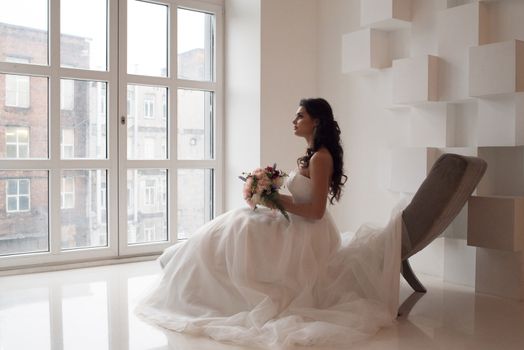 Beautiful bride in her wedding day with flower bouquet looking at window