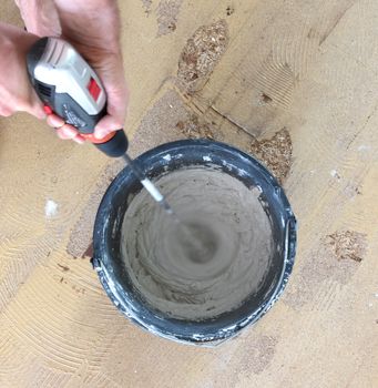 Man is holding drill mixer in his hands. Man is mixing grey concrete and prime in the bucket.