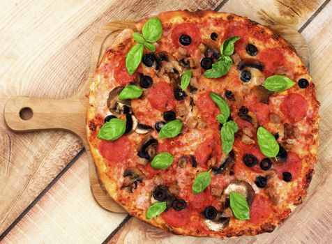 Homemade Pepperoni Pizza with Mushrooms, Black Olives, Ham and Basil on Wooden Cutting Board on Wooden background. Top View