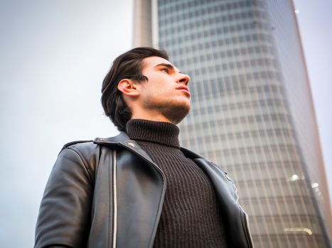 One handsome young man in urban setting in modern city, standing, wearing black leather jacket and jeans