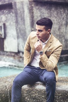 Elegant attractive young man outdoor wearing wool coat, in European city, sitting in front of fountain