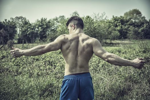 Back of Handsome Muscular Shirtless Young Hunk Man Outdoor in Nature Standing on Grass. Showing Healthy Muscle Body While Looking away