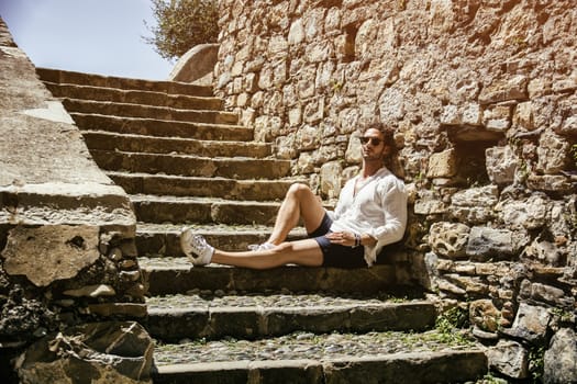 Side view of stylish man in sunglasses sittingon stone stairs and relaxing. Ocean on background.