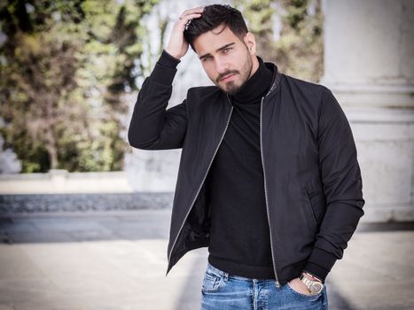 One handsome young man in urban setting in modern city, sitting, wearing black leather jacket and jeans