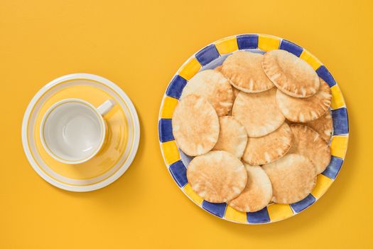 Teacup and colorful plate with mini pita bread on bright yellow background.
