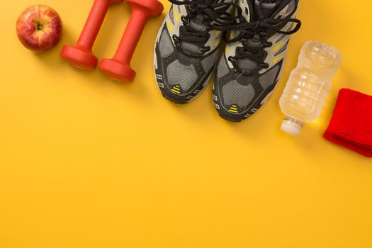 Sport shoes, dumbbells, apple, water bottle and wrist bands. Fitness still life, on yellow background with copy space.