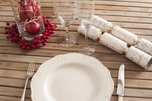 Festive table setting of white plate, cutlery, vine glasses with red berries decorations and wrapped candies over wooden outdoors table surface