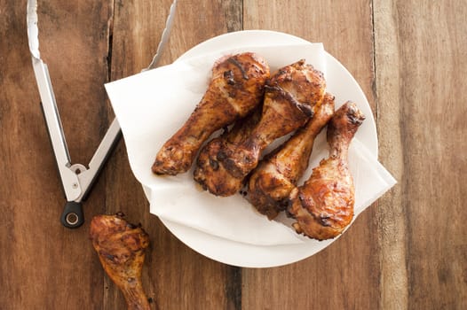 Fried chicken legs on plate with napkin with steel tongs on unpainted wooden table surface background