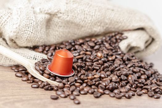 In the foreground a coffee capsule on wooden spoon and   roasted coffee beans with burlap sack on blur wooden background ,close up.