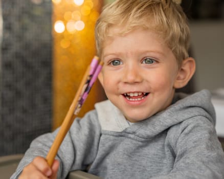 Baby boy sitting in high chair plays with chopsticks and smiling happiness at chinese restaurant.Natural light background with nice bokeh,horizontal photo.