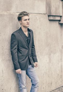 Handsome blond young man, blue eyes, leaning against white, rough stone wall, looking up to a side