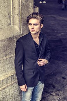 Handsome blond young man, blue eyes, leaning against white, rough stone wall, looking up to a side