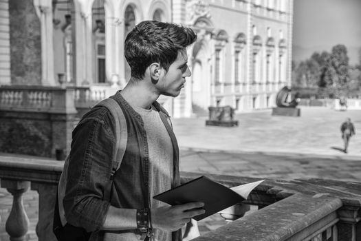 Half Body Shot of a Thoughtful Handsome Young Man, a Tourist, Holding a Guide, Looking Away Outside Historic Building in European City