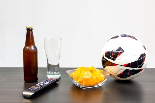 People prepared to watch football on TV with beer. There's beer on the table, ball, TV remote, snacks. Craft beer. Light background.