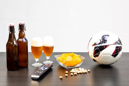 People prepared to watch football on TV with beer. There's beer on the table, ball, TV remote, snacks. Craft beer. Light background.