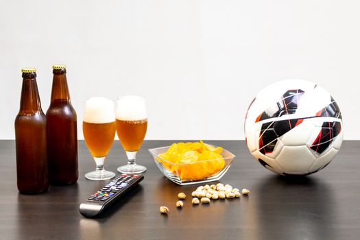 People prepared to watch football on TV with beer. There's beer on the table, ball, TV remote, snacks. Craft beer. Light background.