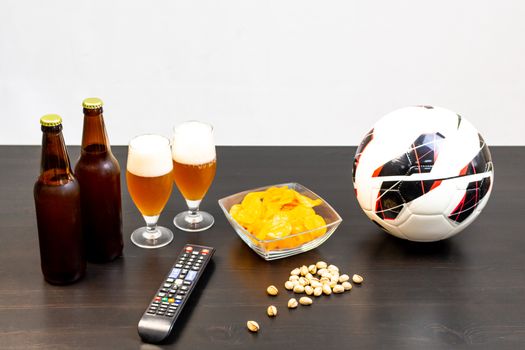 People prepared to watch football on TV with beer. There's beer on the table, ball, TV remote, snacks. Craft beer. Light background.