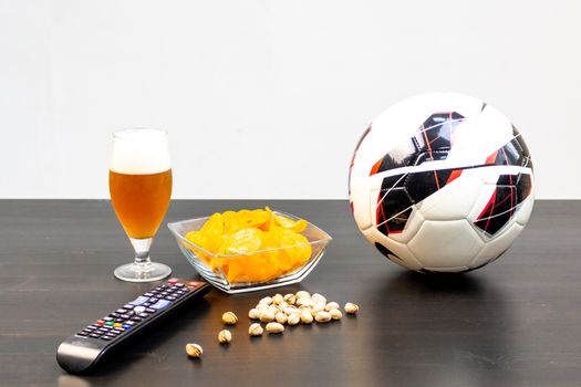 People prepared to watch football on TV with beer. There's beer on the table, ball, TV remote, snacks. Craft beer. Light background.