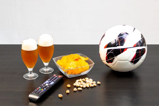 People prepared to watch football on TV with beer. There's beer on the table, ball, TV remote, snacks. Craft beer. Light background.