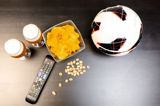People prepared to watch football on TV with beer. There's beer on the table, ball, TV remote, snacks. Craft beer. Light background. The view from the top.