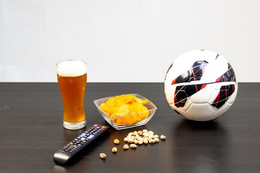 People prepared to watch football on TV with beer. There's beer on the table, ball, TV remote, snacks. Craft beer. Light background.