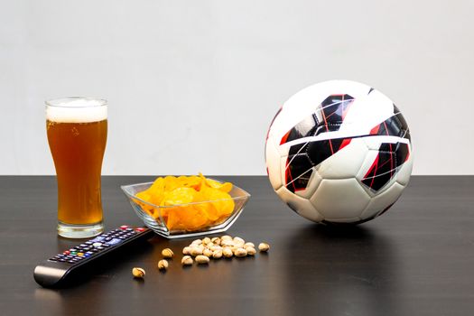 People prepared to watch football on TV with beer. There's beer on the table, ball, TV remote, snacks. Craft beer. Light background.