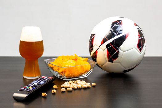 People prepared to watch football on TV with beer. There's beer on the table, ball, TV remote, snacks. Craft beer. Light background.