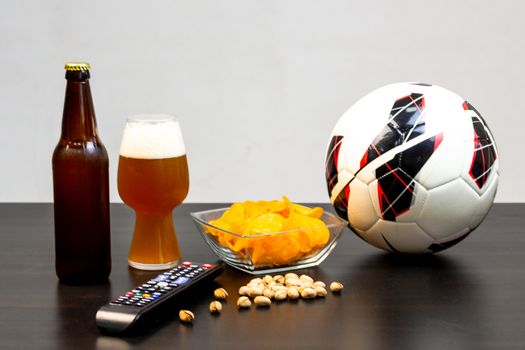 People prepared to watch football on TV with beer. There's beer on the table, ball, TV remote, snacks. Craft beer. Light background.