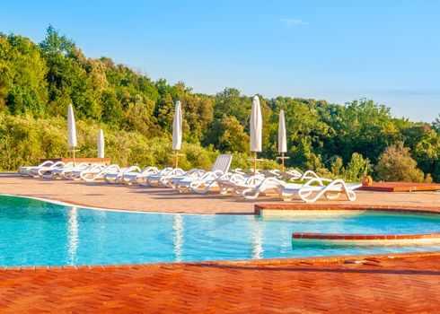 Beautiful luxury swimming pool with bright blue water, umbrellas and sunbeds in Tuscan landscape. Evening summer sunset. Italy.