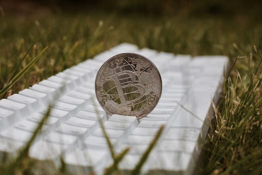 Digital currency physical silver bitcoin coin on white keyboard in green grass.