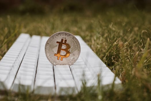 Digital currency physical silver bitcoin coin on white keyboard in green grass.