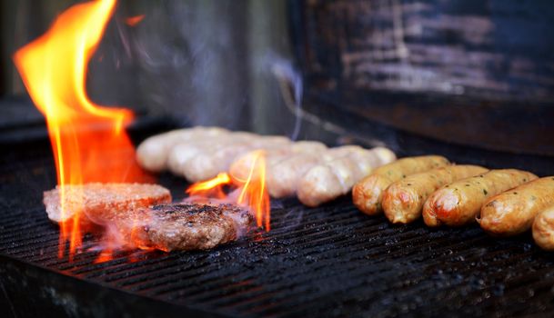 Flame rises around a hamburger on a barbecue grill, with sausages cooking in the background
