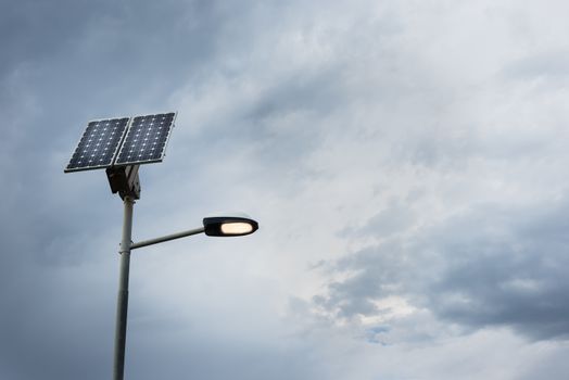 Solar panel on street lamp post with light on and cloudy sky.