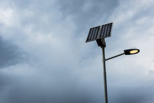 Solar panel on street lamp post with light on and cloudy sky.