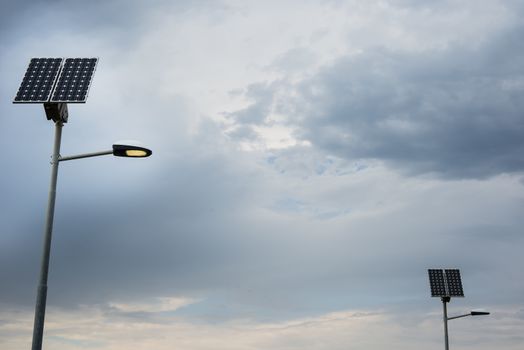 Solar panel on street lamp post with light on and cloudy sky.