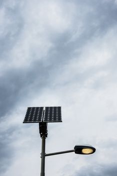 Solar panel on street lamp post with light on and cloudy sky.