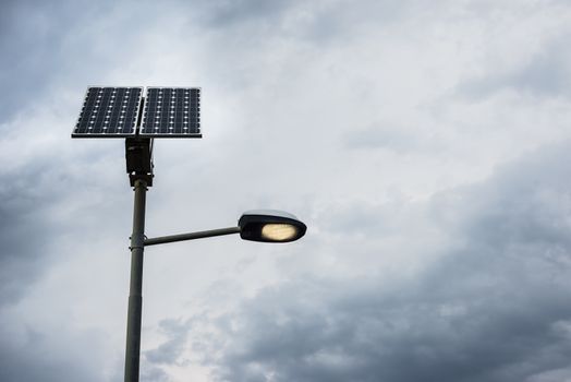 Solar panel on street lamp post with light on and cloudy sky.