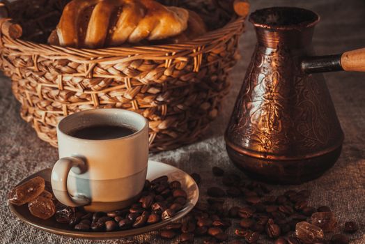 A cup of coffee on textile with coffee beans, dark candy sugar, pots, basket and cake