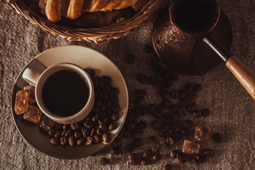 A cup of coffee on textile with coffee beans, dark candy sugar, pots, basket and cake