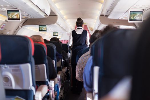 Interior of commercial airplane with passengers on seats during flight. Stewardess in dark blue uniform walking the aisle. Horizontal composition.