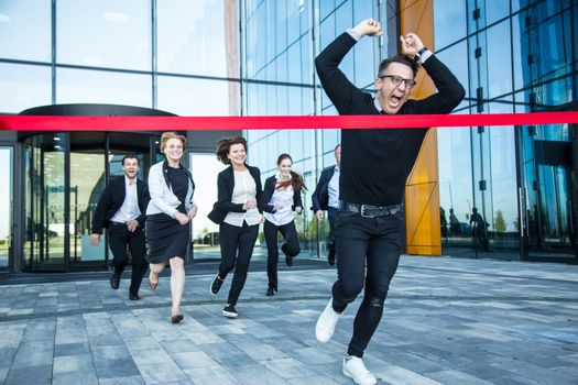 Group of happy business people running from office building crossing red ribbon finish line