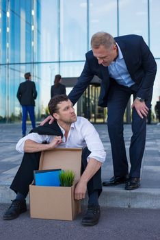 Fired business man sitting frustrated and upset on the street near office building with box of his belongings. He lost work. Other businessman comforts and encourages him