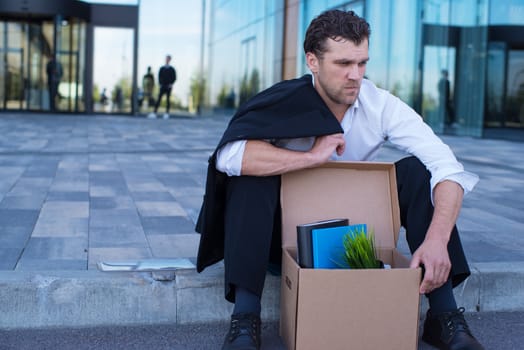 Fired business man sitting frustrated and upset on the street near office building with box of his belongings. He lost work