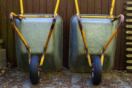 Two wheelbarrows standing in front of wooden wall outside.