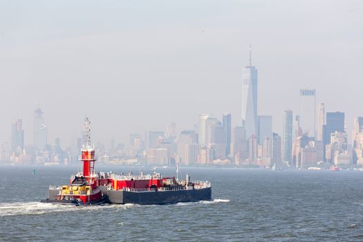 Freight tug pushing cargo ship to the port in New York City and Lower Manhattan skyscarpers skyline in background. New York City, USA.