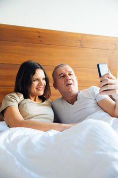 Attractive and Happy Mid Aged Couple Enjoying in the Bed in the Morning and Using a Smartphone and Taking Selfie Photo