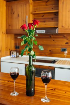 Three Red Roses In Bottle And Two Glasses Of Red Wine On Wooden Table